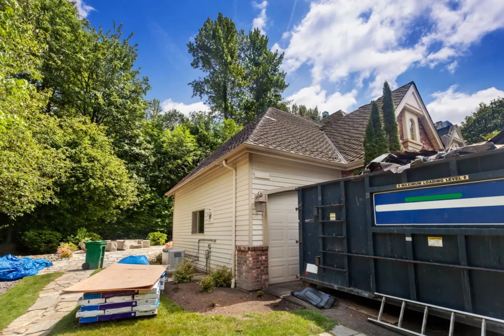 Suburban home in the middle of a re-roofing project, with new shingles and plywood waiting to be installed and dumpster to contain the debris.
